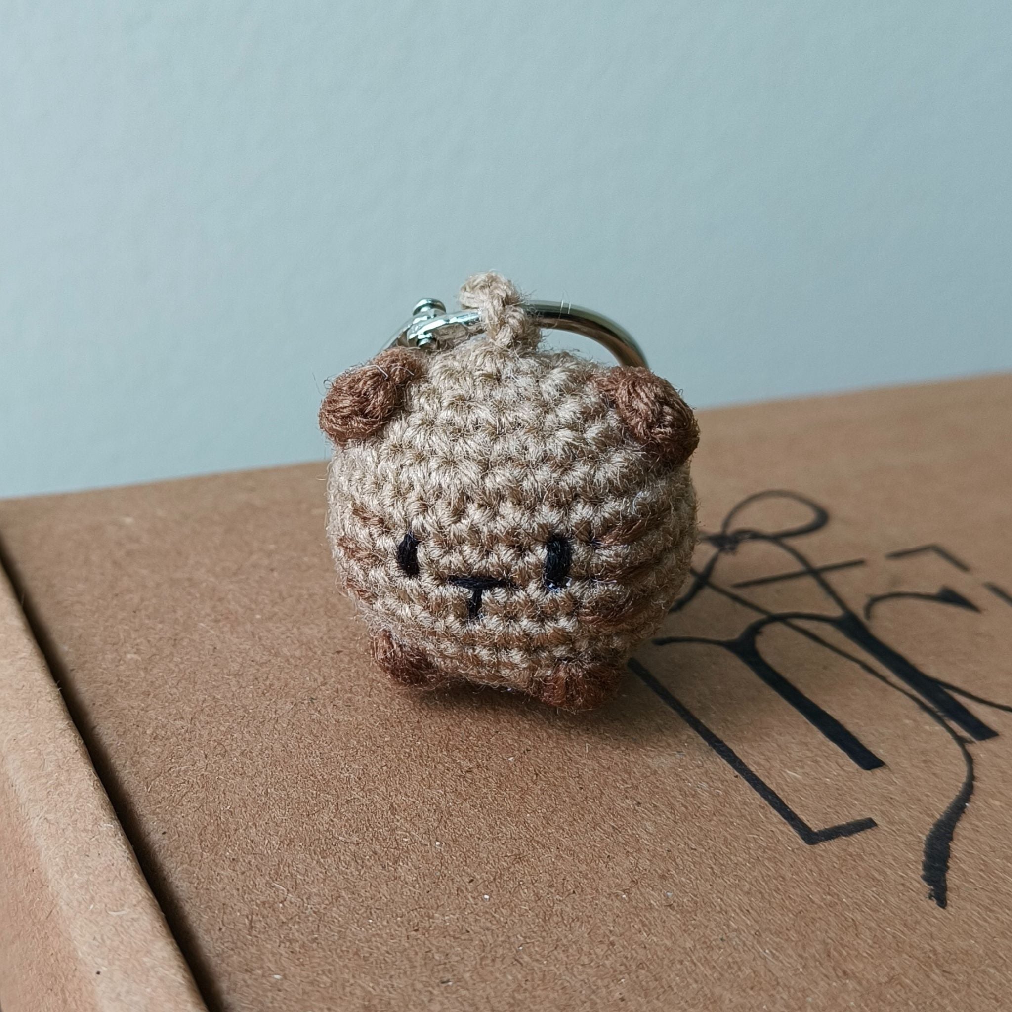 Small crocheted brown and beige keychain on a cardboard surface with a light blue background
