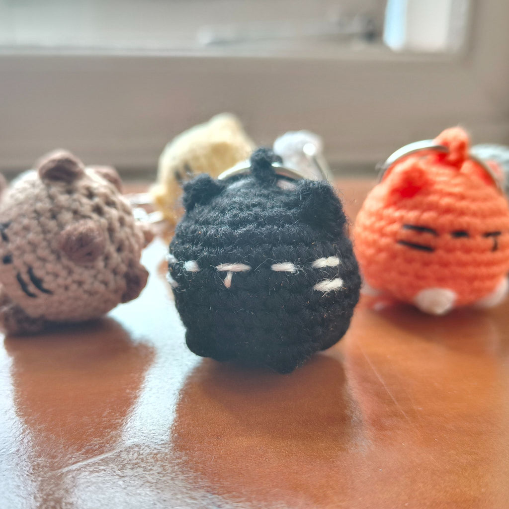 Small crocheted black cat-themed keychain in focus with other kitty keychains in background on a wooden surface.