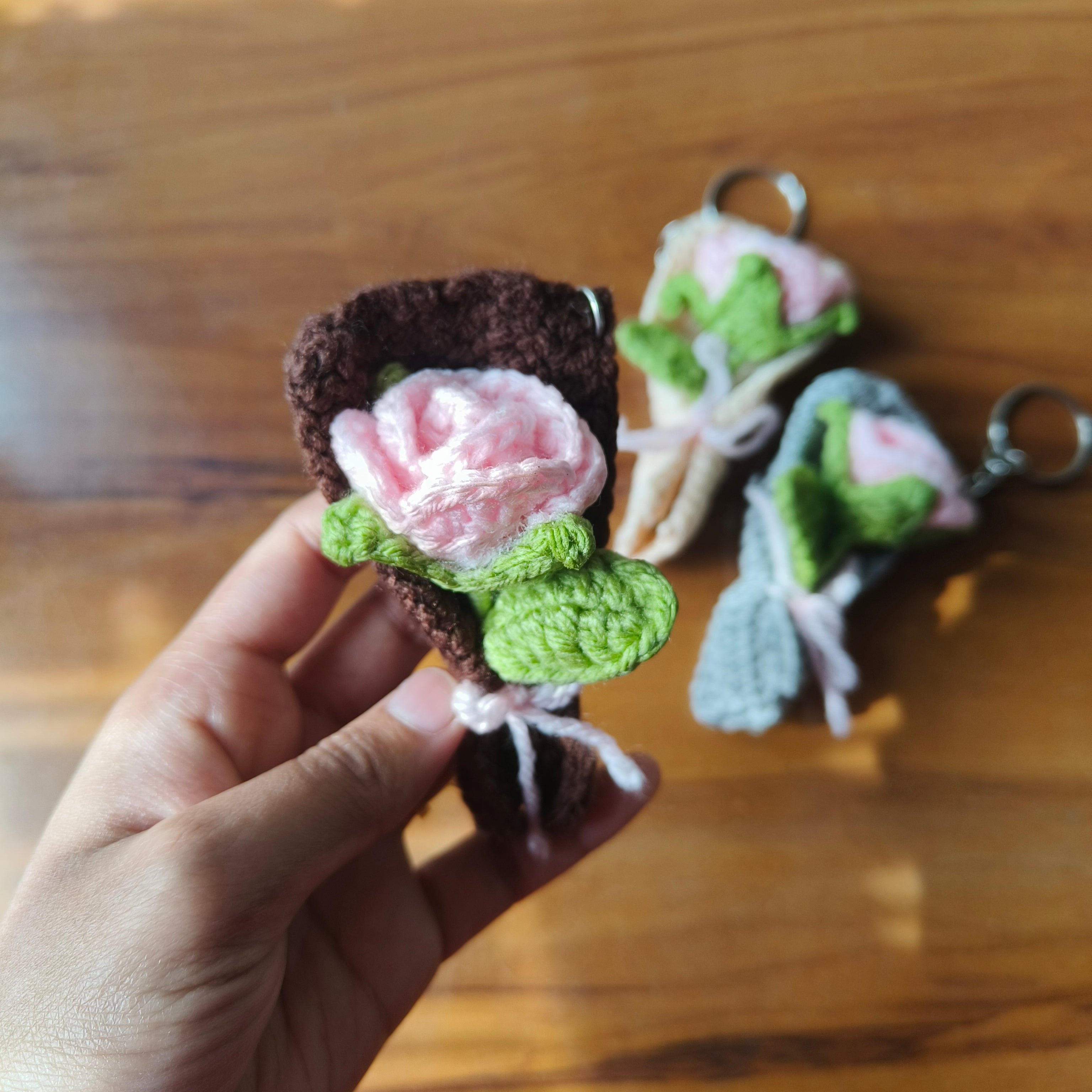 Hand holding a crocheted flower keychain with two more keychains in the background on a wooden surface.