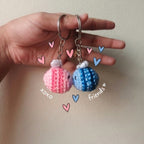 Hand holding two small bee crocheted keychains shaped like balloons, one pink and one blue, with decorative hearts and text on a plain background.