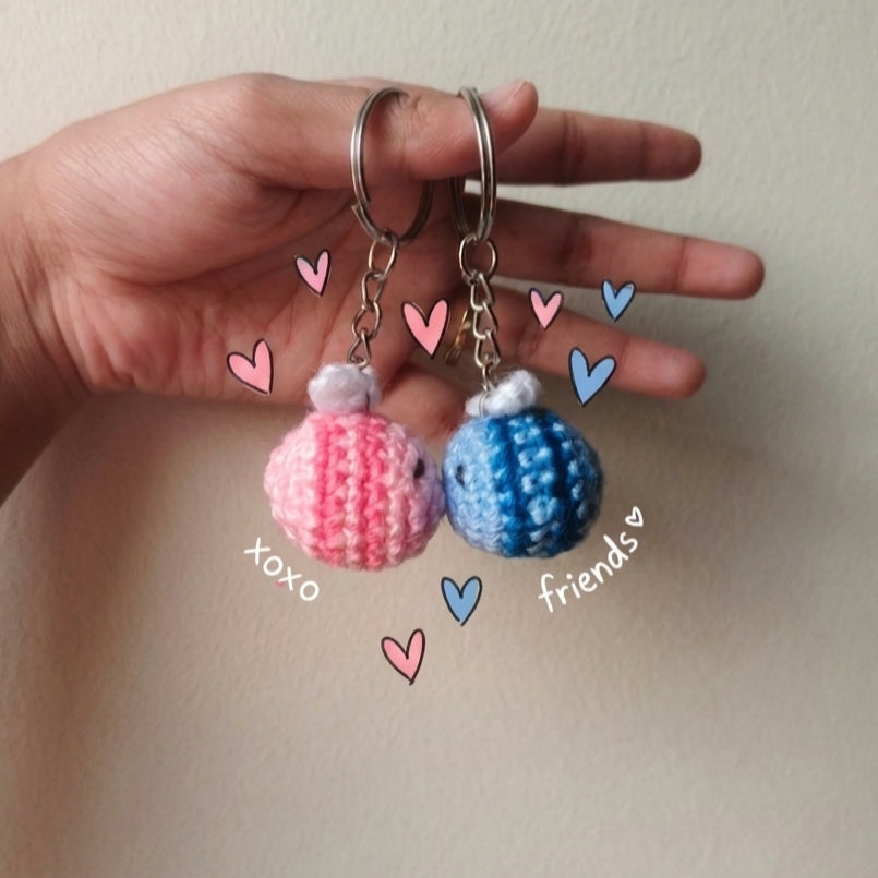 Hand holding two small bee crocheted keychains shaped like balloons, one pink and one blue, with decorative hearts and text on a plain background.